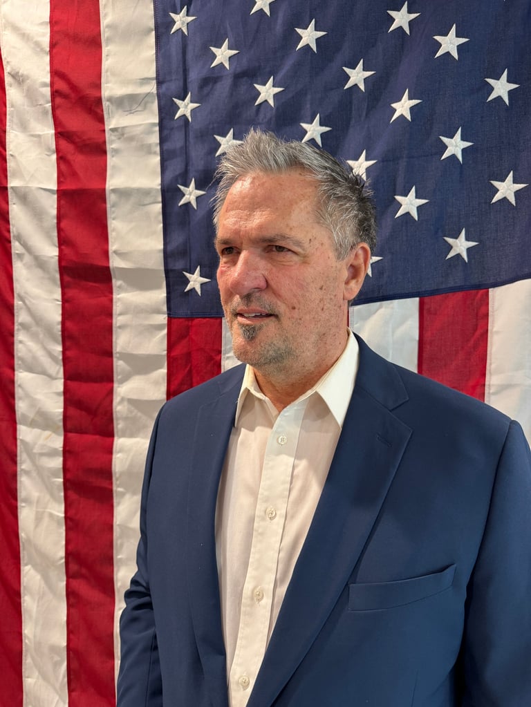 Middle-aged man in dark blazer and white shirt posing in front of American flag background