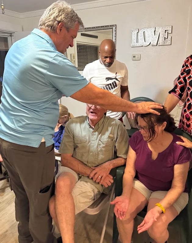 A group of people gather indoors, with several standing around two seated individuals during the Baptize the Repentant event