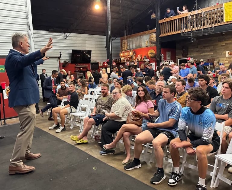 Man speaking to a large seated crowd in an indoor warehouse or fitness facility with industrial architecture and staging visible