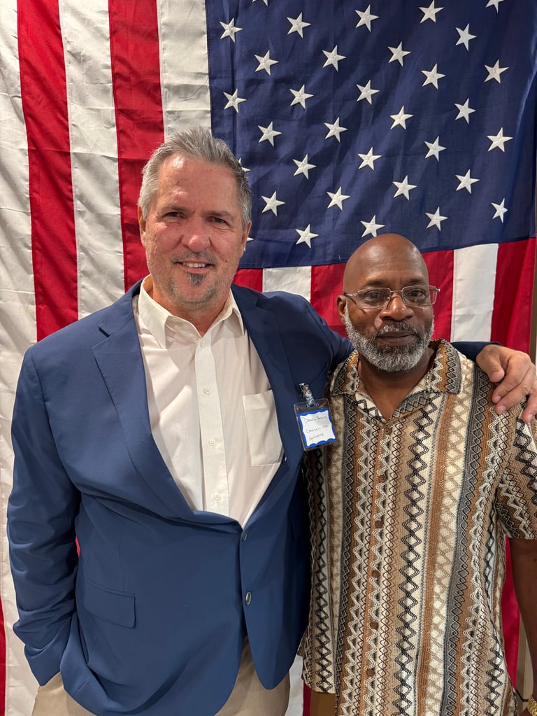 Two men smiling at camera in front of American flag. Man on left wears navy blazer and white shirt. Man on right wears patterned cardigan.